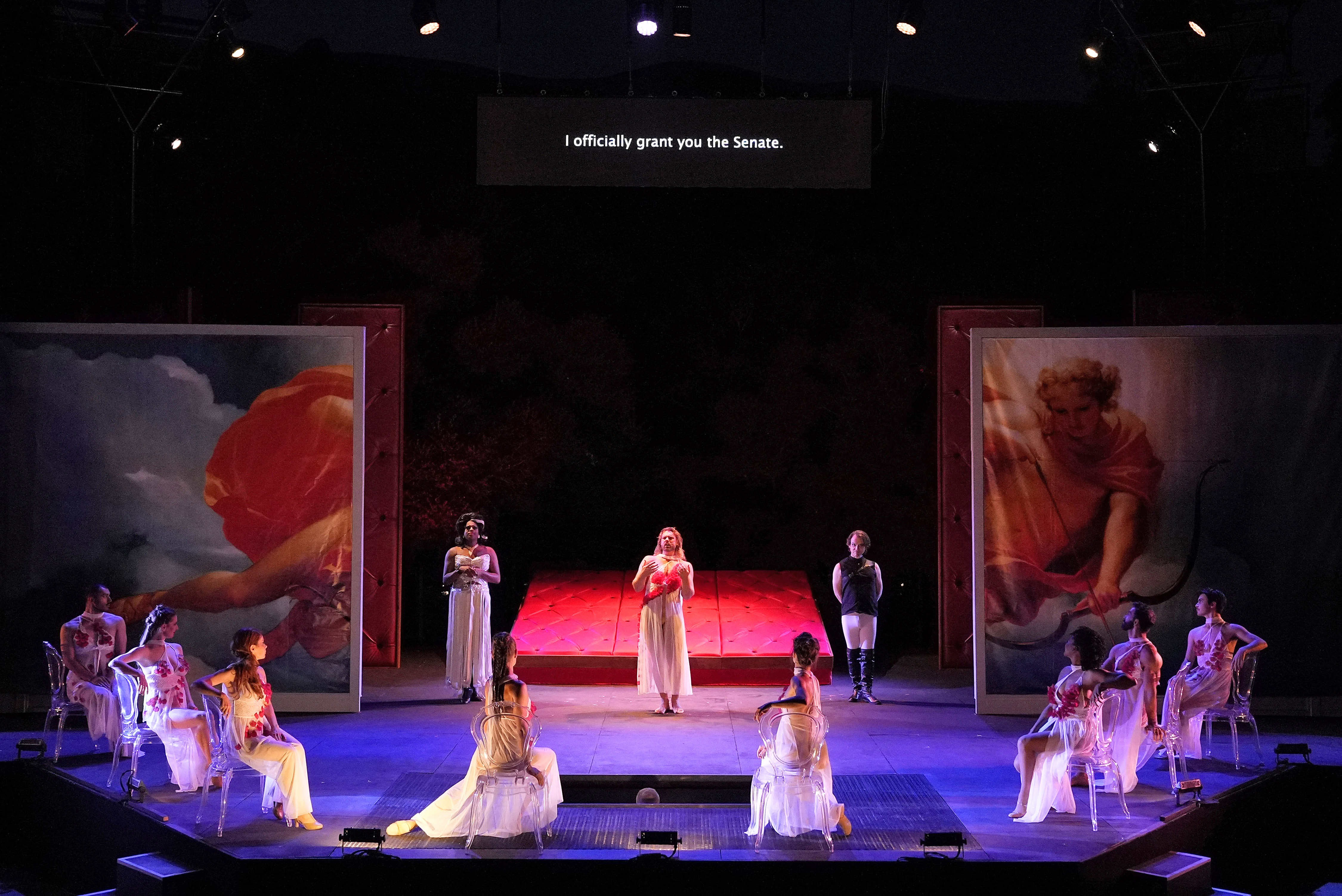 A ring of feminine dancers sit in chairs, attentively looking at the emperor in the middle.